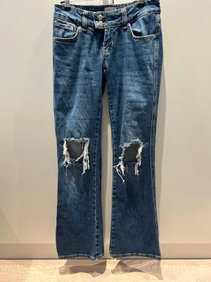 Blå slitna bootcut jeans - Säljer ett par blå bootcut jeans med slitna hål på båda knäna och klassiska fem fickor. Jeansen har en cool tvätt och råa kanter vid hålen för en trendig look. Jeansen är lågmidjade