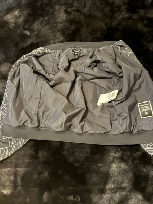 Armani jacka    Size S - Snygg svart bomberjacka från Emporio Armani med ett unikt grafiskt mönster i vitt över hela jackan. Jackan har ribbade muddar, dragkedja framtill och två sidofickor. Tillverkad i ett mjukt och bekvämt material som passar perfekt till streetwear-stilen.