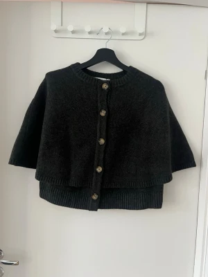 Mörkgrå ullcape med knappar - Stilren mörkgrå cape i ullmix med stora knappar framtill och ribbade kanter. Modellen har en avslappnad passform och trekvartsärmar, perfekt för lager på lager och en cool look.