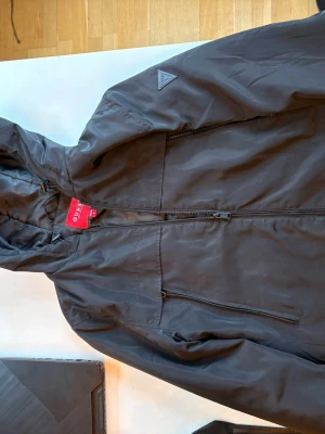 Svart vindjacka från Guess - Snygg svart vindjacka från Guess med huva och dragkedja framtill. Jackan har en bröstficka med dragkedja och diskret Guess-logga på axeln. Tillverkad i lätt och slitstarkt material som passar perfekt för blåsiga dagar.