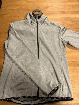 Grå zip-hoodie från CRW  - Snygg grå zip-hoodie från CRW med svart dragkedja och huva. Tröjan har svarta detaljer med Cru-logga längs nederkant och ärmslut. Perfekt för träning. Två fickor framtill och bekväm passform.