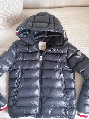 Moncler alberic giubbotto 164 cm - Snygg marinblå dunjacka från Moncler med glansig finish, huva och två dragkedjefickor framtill. Jackan har Moncler-logga på ärmen och coola rödvita detaljer på huva och ärmslut. Insidan har ett unikt serietryck. Perfekt för kalla vinterdagar.