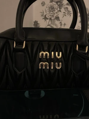 Svart handväska från Miu Miu - Snygg svart handväska från Miu Miu i quiltat läder med guldfärgade detaljer och logotyp framtill. Väskan har dubbla handtag och dragkedja upptill samt innerfack med dragkedja. Perfekt för dig som gillar lyxiga accessoarer med stilren design.