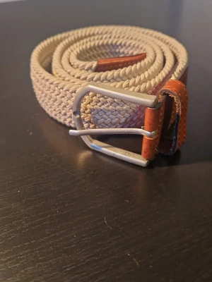 Beige flätat bälte med skinn-detaljer - Snyggt beige flätat bälte med bruna skinn-detaljer och silverfärgat spänne. Perfekt accessoar för att lyfta din outfit med en stilren touch. Bältet har en klassisk design och passar till många olika looks.