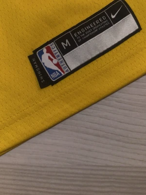 Gul Lakers basketlinne Nike NBA barn 10-12 år - Säljer en gul basketlinne från Nike med Lakers-tryck och nummer 6 på bröstet. Linnet är officiellt NBA Authentics Swingman och har lila och vita detaljer runt ärmar och hals. Materialet är lätt och andas, perfekt för basket eller träning.