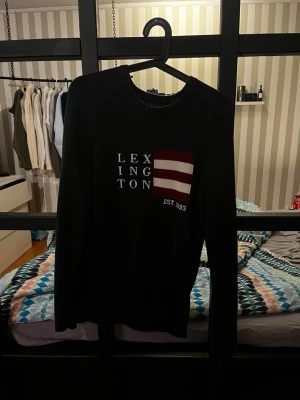 Lexington stickad tröja - Svart stickad tröja från Lexington med vit text och en röd-vit randig detalj på bröstet samt 'EST. 1993'. Tröjan har rund halsringning och långa ärmar. Perfekt för dig som gillar klassisk och stilren design.