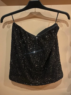 Svart glittrig tubtopp - Snygg svart tubtopp täckt av glittriga paljetter.       Bygeln har jag klippt o tagit ur men inget som syns. Det är silikon rand runt brösten så den glider ej ner