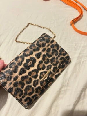 Kuvertväska med leopardmönster - Snygg kuvertväska med leopardmönster och guldfärgad kedja. Väskan har en rektangulär form och stängs med ett magnetlås. Materialet är konstläder med struktur och detaljer i guld. Perfekt för att lyfta din outfit med lite extra attityd.