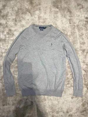 Grå tröja från Polo Ralph Lauren - Snygg grå tröja från Polo Ralph Lauren med rund halsringning och diskret broderad logga på bröstet. Tröjan har långa ärmar och ribbade muddar vid ärmslut och nederkant. Perfekt för en stilren och avslappnad look.  8/10 ny pris c 1500 