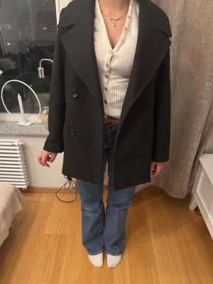 Svart dubbelknäppt rock med bred krage - Stilren svart rock med dubbelknäppning och bred krage. Klassisk modell som går till låret och har fyra knappar framtill. Perfekt för dig som gillar en tidlös och enkel look. Materialet ser ut att vara ullblandning.