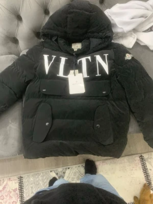 Svart dunjacka Moncler x Valentino - Svart dunjacka med stor vit VLTN-logga framtill och Moncler-märke på ärmen. Jackan har huva, stora fickor med lock och knappar samt en quiltad design. Perfekt för kalla dagar och riktigt snygg streetstil.