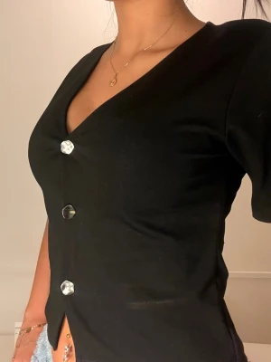 Svart v-ringad topp med knappar - Snygg svart topp med v-ringning och korta ärmar. Toppen har dekorativa knappar framtill och en croppad passform som ger en trendig look. Perfekt att matcha med jeans eller kjol för en stilren outfit.