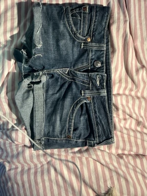 Blå jeansshorts från True Religion - Säljer ett par blå jeansshorts från True Religion med slitna detaljer och råa kanter. Shortsen har vita kontrastsömmar, klassiska fickor fram och bak samt dekorativa knappar på bakfickorna. Perfekta för sommaren!