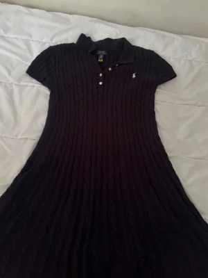 Mörkblå stickad klänning från Polo Ralph Lauren - Mörkblå kabelstickad klänning från Polo Ralph Lauren i barnstorlek XL. Klassisk modell med vit broderad logga på bröstet och det ikoniska kabelstickade tyget. (Original därför är priset högt.)