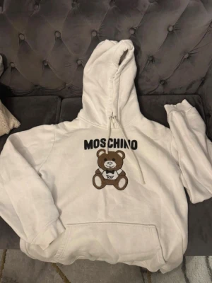 Vit Moschino hoodie med nalletryck - Vit hoodie från Moschino med stor huva och dragsko. På bröstet finns ett broderat tryck av en brun nalle med texten 'TOY' samt Moschino-loggan i svart ovanför. Hoodien har en klassisk känguruficka och ribbade muddar. Perfekt för en trendig och avslappnad stil.