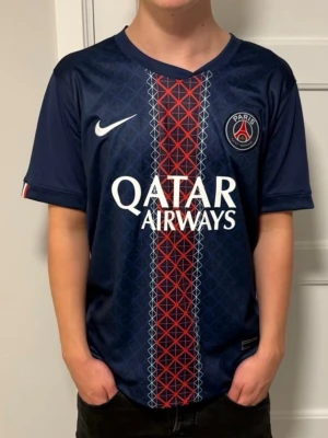 PSG matchtröja blå Nike - Snygg blå PSG matchtröja från Nike med röd och vit detalj i mitten, klubbmärke på bröstet och Qatar Airways som sponsor. Tillverkad i lätt och ventilerande material, perfekt för fotboll eller träning. Korta ärmar och klassisk passform.