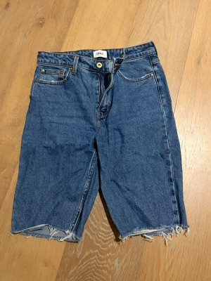 Blå jeansshorts från ONLY - Snygga blå jeansshorts från ONLY med råa, fransiga benslut och klassisk femficksdesign. Shortsen har normal passform och stängs med knapp och dragkedja. Perfekta för en avslappnad stil under varma dagar.