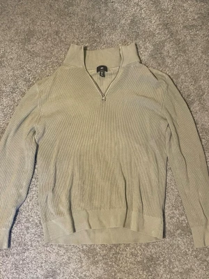 Beige stickad tröja från H&M - Säljer en beige stickad tröja från H&M i slim fit-modell. Tröjan har halv dragkedja framtill, hög krage och ribbstickad struktur. Perfekt för dig som gillar stilrena och enkla plagg med en modern touch.
