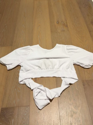 Vit croppad topp med knyt från H&M - Trendig vit croppad topp från H&M med puffiga korta ärmar och breda knytband i midjan. Toppen har en ribbad struktur och rund halsringning. Perfekt för dig som vill ha en cool och stilren look.
