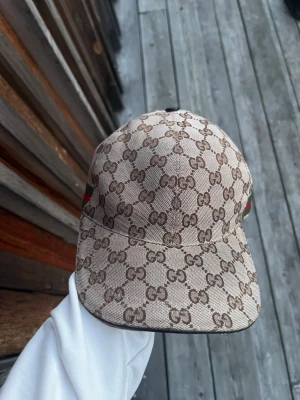Gucci keps med GG-mönster - Snygg beige keps från Gucci med klassiskt GG-monogram över hela kepsen och grönt/rött band på sidan. Kepsen har böjd skärm och är tillverkad i canvasmaterial. Perfekt för dig som vill sticka ut med en lyxig accessoar.