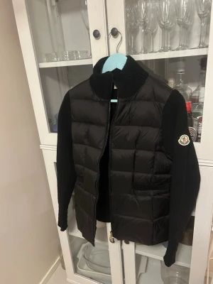 Svart cardigan från Moncler - Snygg svart cardigan från Moncler med stickade ärmar och hög krage. Jackan har quiltad front och rygg i glansigt material, samt ribbstickade muddar. Moncler-logga på vänster ärm. Perfekt för kalla vinterdagar och riktigt trendig look. Passar typ männens M