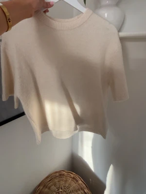 Beige stickad kortärmad tröja - Mysig beige stickad tröja med korta ärmar och rund hals. Tröjan har en enkel och stilren design med ribbade kanter vid hals, ärmslut och nederkant. Perfekt för lager på lager och en avslappnad look.