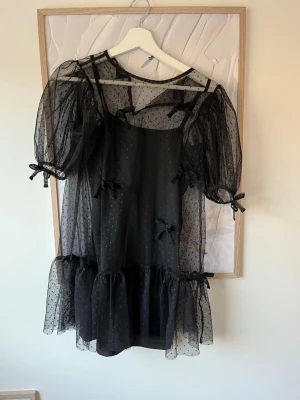 Svart transparent kortklänning med rosetter - Säljer en svart kortklänning i transparent mesh med små prickar, puffärmar och söta rosetter på ärmar och fram. Klänningen har volangkant nedtill och en loose passform. Perfekt för dig som gillar en lekfull och trendig stil.