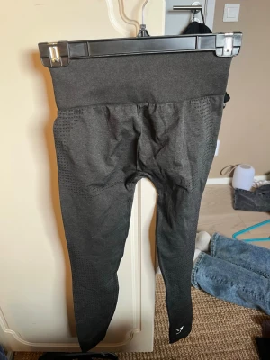 Svarta leggings Gymshark - Svarta leggings från Gymshark med hög midja och diskret mönster längs benen. Tillverkade i stretchigt syntetmaterial som sitter tight och följsamt. Gymshark-logga på höften. Perfekta för både träning och chill.