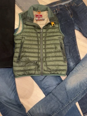 Grön dunväst från Parajumpers - Snygg grön dunväst från Parajumpers med dragkedja framtill och två sidofickor med dragkedja. Västen har en hög krage och ikonisk gul detalj vid halsen. Lätt vadderad och perfekt för lager-på-lager-stil. Passar till jeans och streetwear.