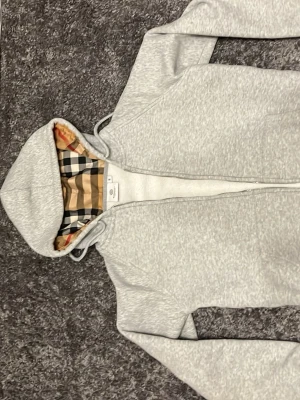 Grå hoodie från Burberry med huva passar 158-164/xs - Snygg grå zip-hoodie från Burberry med klassiskt rutigt foder i huvan. Tillverkad i mjukt material och har dragsko i huvan samt fickor framtill. Perfekt för dig som gillar stilrena plagg med exklusiv känsla.