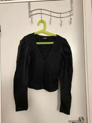 Svart blus med puffärmar från Gina Tricot - Trendig svart blus från Gina Tricot med smockad kropp, djup V-ringning och puffiga långärmar. Knäppning framtill och figurnära passform ger en snygg siluett. Perfekt att styla med både jeans och kjol. Materialet är en mix av bomull och elastan.