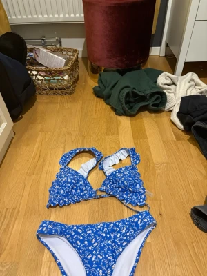Blå bikini med blommönster - Söt blå bikini med vitt småblommigt mönster och volangdetaljer på överdelen. Bikinin har vadderade kupor och breda axelband för extra stöd. Underdelen har klassisk passform och samma blommiga design. Perfekt för strandhäng eller poolparty!