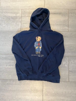 Marinblå hoodie Polo Bear Ralph Lauren - Snygg marinblå hoodie från Ralph Lauren med den ikoniska Polo Bear-trycket på bröstet. Klassisk huva med dragsko och ribbade muddar. Perfekt för dig som gillar streetwear och vill ha en avslappnad men stilren look.