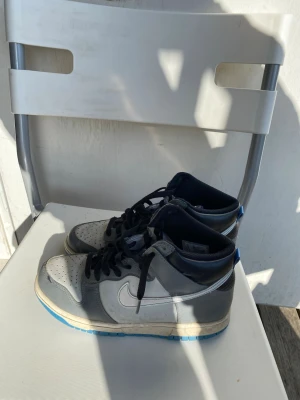 Nike grå/blå high sneakers - Nike sneakers i high-modell med grå och vita paneler, svarta skosnören och blå detaljer på plösen och yttersulan. Klassisk swoosh på sidan och perforerad tåbox för extra ventilation. Snygg retrostil som passar till streetwear.
