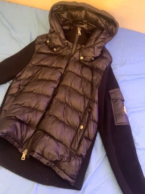 Svart dunväst med huva från Moncler - Snygg svart dunväst från Moncler med stickade ärmar och stor huva. Västen har dragkedja framtill, två fickor med tryckknappar och en Moncler-logga på ärmen. Materialet är en mix av glansigt dun och ribbstickad ull för extra stilpoäng.