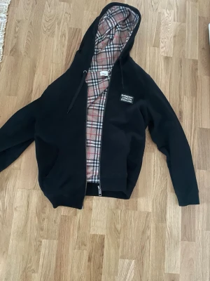 Svart Burberry hoodie med rutigt foder - Snygg svart hoodie från Burberry med dragkedja och klassiskt rutigt foder i beige, rött och svart. Märket syns tydligt på bröstet och insidan. Hoodien har huva med snörning och är perfekt för dig som vill ha en exklusiv streetwear-look.