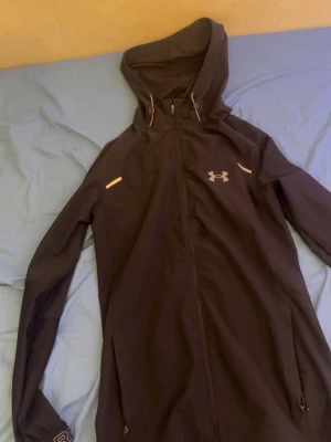 Svart vindjacka från Under Armour - Snygg svart vindjacka från Under Armour med huva och hel dragkedja. Jackan har reflekterande detaljer på bröstet och ärmarna samt två fickor med dragkedja. Perfekt för blåsiga dagar och sportiga outfits.