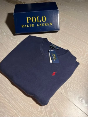 Mörkblå tröja från Polo Ralph Lauren - Mörkblå tröja från Polo Ralph Lauren med klassisk V-ringning och röd broderad logga på bröstet. Tröjan är i mjukt material och har en stilren, tidlös design som passar perfekt till jeans eller chinos.box ingår inte men tagen/prislappen ingår