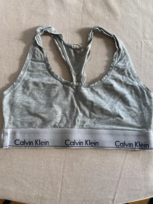 Grå sport-bh från Calvin Klein - Grå sport-bh från Calvin Klein med brottarrygg och bred resår med logga runt om. Mjuk och stretchig bomullsblandning som ger en bekväm passform. Perfekt för en avslappnad och sportig stil.
