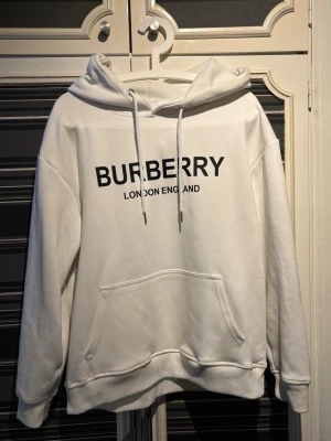 Vit hoodie från Burberry - Snygg vit hoodie från Burberry med svart logga och texten 'London England' på bröstet. Hoodien har stor huva med en klassisk magficka framtill. Priset kan diskuteras.