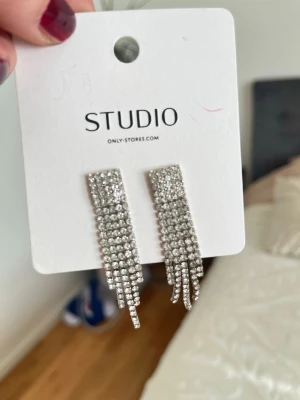 Glittriga hängande örhängen Studio - Snygga silverfärgade örhängen från Studio med flera rader av gnistrande stenar som hänger ner i en vågig form. Perfekta för att ge extra bling till din look. Lätta att bära och riktigt trendiga.