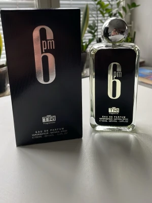 6pm Eau de Parfum från TRI Fragrances - Snygg parfymflaska från TRI Fragrances med namnet 6pm. Flaskan är genomskinlig med svart etikett och silverdetaljer, och har en rund silverfärgad kork. Förpackningen är svart med stilren design. Innehåller 100 ml Eau de Parfum.
