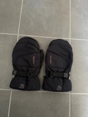 Svarta Hestra tumvantar med spänne, stl 10 - Svarta tumvantar från Hestra med justerbart spänne runt handleden och diskret logga på mudden. Vantarna är vadderade och har en enkel, sportig design som passar perfekt för kalla dagar.