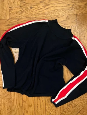 Svart långärmad topp med ränder - Snygg svart långärmad topp med röda och vita ränder längs ärmarna. Hög hals och sportig look, perfekt till jeans eller kjol. Materialet känns mjukt och stretchigt.