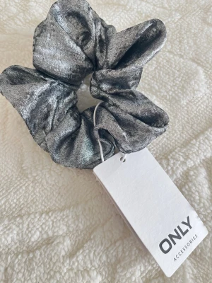 Metallic silver scrunchie från ONLY - Säljer en snygg scrunchie i metallic silver från ONLY. Hårsnodden har en glansig finish och är tillverkad i ett mjukt tyg som ger volym och en trendig look. Perfekt för att piffa upp din frisyr med lite extra glans.