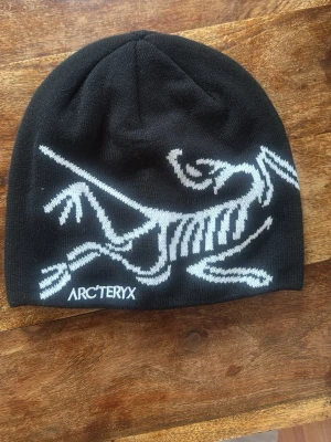 Svart mössa från Arc'teryx - Säljer en svart mössa från Arc'teryx med stor vit logga i form av ett skelettmönster på framsidan. Mössan är stickad och har en klassisk, rundad form som sitter skönt på huvudet. Perfekt för dig som gillar stilrena accessoarer med cool design.