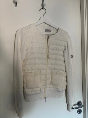 Vit cardiganjacka från Moncler - Snygg vit cardiganjacka från Moncler med quiltad front, två fickor med knappar och dragkedja framtill. Ärmarna är stickade och har Moncler-logga på vänster ärm. Perfekt för lager på lager och en clean look. Kan tänka mig att gå ner i pris så buda gärna! Nypris ca 9000