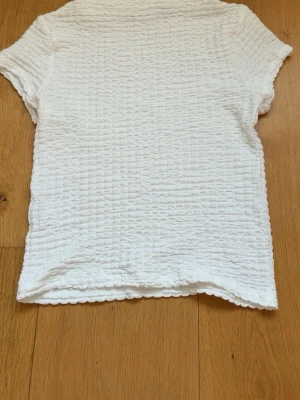 Vit strukturerad topp från Kappahl - Vit topp från Kappahl med korta ärmar och strukturerat tyg som ger en cool och modern känsla. T-shirten har en lätt vågig kant vid halsen och nederkanten. Perfekt för dig som gillar en enkel men ändå unik stil.