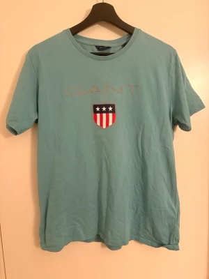 Ljusblå GANT t-shirt - Säljer en ljusblå t-shirt från GANT med klassiskt tryck framtill och ett sköldmärke med stjärnor. Den är storlek 176cm i barn storlek alltså XS-S i vuxen storlek. Den har endast ett jätte litet hål men annars är den jätte gott skick.