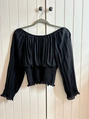 Svart blus - Trendig svart off shoulder topp eller vanlig blus med långa ärmar i plisserat tyg. Smockad midja och ärmslut ger en snygg passform och volangdetaljer längst ner. Materialet känns lätt och flowy. Storlek S men passar även XS 